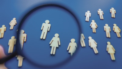 Attēlā uz zila fona redzamas koka vīriešu un sieviešu figūriņas, priekšplānā redzama lupa, kas palielina atsevišķas figūriņas