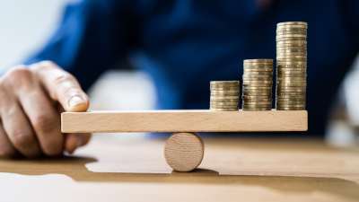 Attēlā redzama cilvēka roka, kura tur līdzsvarā tur trīs monētu kaudzītes uz līdzsvara šūpolēm