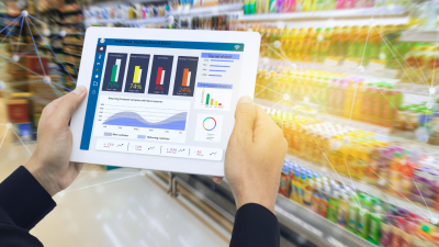 Attēlā tuvplānā redzama planšete, kurā redzami dažādi dati, fonā produktu plaukti kādā tirdzniecības vietā