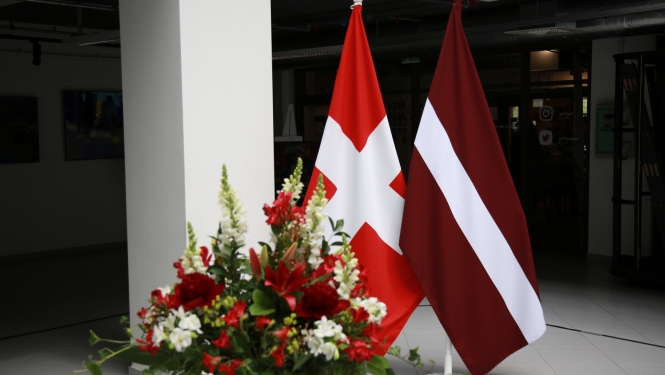 Latvijas un Šveices karogi uz statīviem, priekšplānā ziedu pušķis